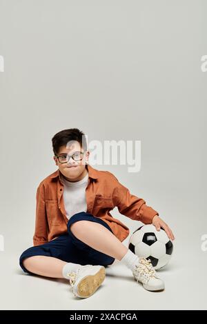 Kleiner Junge mit Down-Syndrom mit Brille sitzt auf dem Boden mit Fußball. Stockfoto