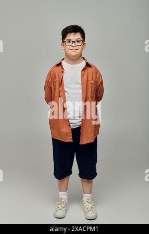 Kleines Kind mit Down-Syndrom in Brille und Shorts, selbstbewusst vor grauem Hintergrund. Stockfoto