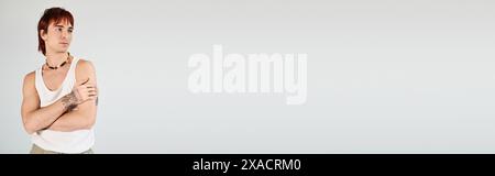 Ein stilvoller junger Mann posiert selbstbewusst in einem weißen Tanktop und steht mit gekreuzten Armen in einem Studio mit grauem Hintergrund. Stockfoto