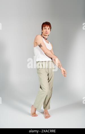 Ein stilvoller junger Mann posiert in einem Studio vor grauem Hintergrund, trägt ein weißes Tanktop und khakifarbene Hosen. Stockfoto