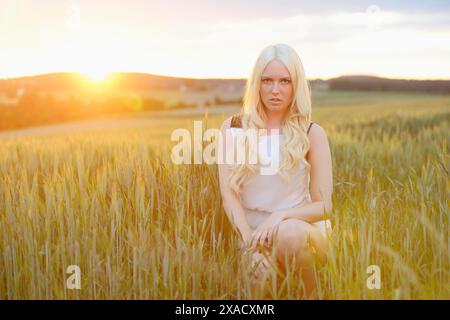 Eine blonde Frau in einem weißen Kleid sitzt während des Sonnenuntergangs auf einem grasbewachsenen Feld Stockfoto