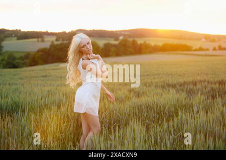 Eine blonde Frau steht auf einem Feld bei Sonnenuntergang, in einem weißen Kleid und umgeben von warmem Licht Stockfoto