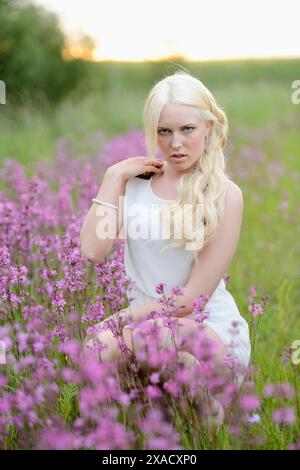 Eine blonde Frau in einem weißen Kleid sitzt nachmittags zwischen violetten Blumen auf einem natürlichen Feld Stockfoto