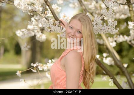 Frau mit blonden Haaren in einem rosa Kleid lächelt, während sie sich an einen Kirschblütenbaum in einer sonnigen Umgebung im Freien lehnt Stockfoto