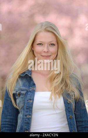 Frau mit blondem Haar, die eine Jeansjacke und ein weißes Oberteil trägt, lächelt mit einem weichen rosa Hintergrund im Freien Stockfoto