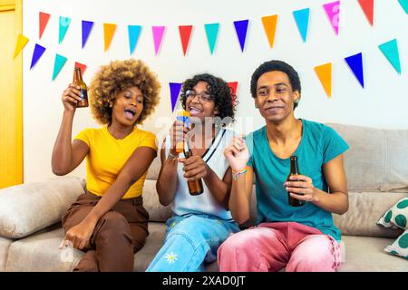 Verschiedene Freunde singen und trinken Bier während der Karaoke zu Hause, sitzen auf dem Sofa mit Party-bunten Girlanden an der Wand Stockfoto