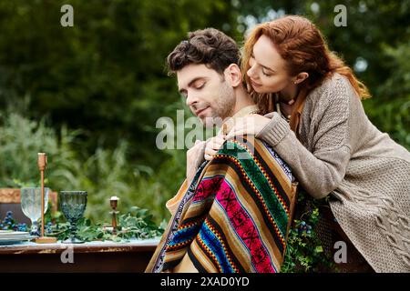 Ein Mann und eine Frau in Boho-Kleidung sitzen an einem mit Decken bedeckten Tisch in einem grünen Park und genießen ein gemütliches und romantisches Date. Stockfoto