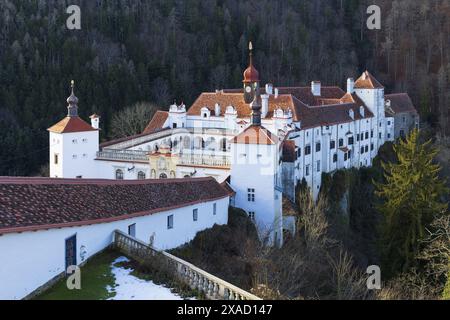 Fotografie eines alten weißen Barockschlosses mit rotem Ziegeldach, umgeben von alpinen Bergen am sonnigen Wintertag Stockfoto