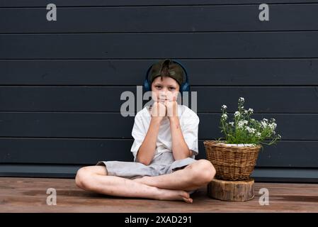 Geballter Jugendlicher in Kopfhörern, der auf einer Holzterrasse in der Nähe einer Blume sitzt Stockfoto