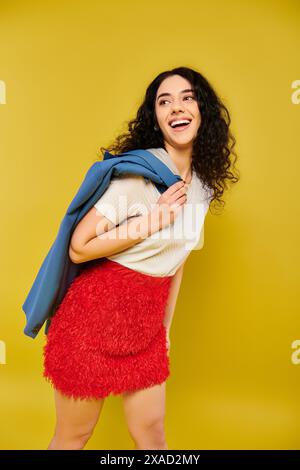 Eine junge Frau mit lockigen Haaren posiert in einem leuchtenden roten Rock und weißem Hemd und zeigt ihre Emotionen vor gelbem Hintergrund. Stockfoto