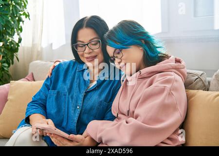 Asiatische Mutter und Tochter in legeren Kleidern sitzen auf einer Couch, konzentrieren sich auf einen Handybildschirm, verbinden sich und verbringen wertvolle Zeit miteinander. Stockfoto