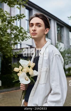 Ein junger, stilvoll gekleideter Mann hält stolz einen Blumenstrauß vor einem großen Gebäude. Stockfoto