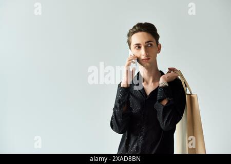 Eine junge queere Person hält Einkaufstaschen, während sie vor grauem Studiohintergrund am Telefon plaudert. Stockfoto