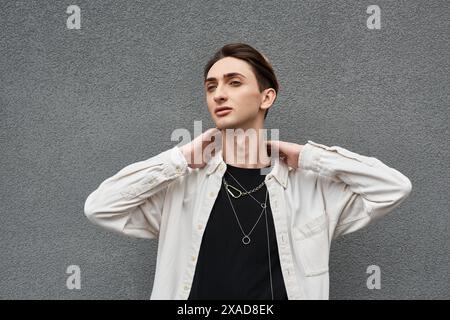 Eine junge queere Person posiert selbstbewusst vor einer grauen Wand und zeigt seine stilvolle Kleidung mit Stolz und Selbstvertrauen. Stockfoto