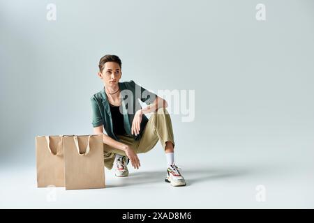 Eine junge queere Person, die auf dem Boden sitzt, umgeben von bunten Einkaufstaschen in einem Studio auf grauem Hintergrund. Stockfoto