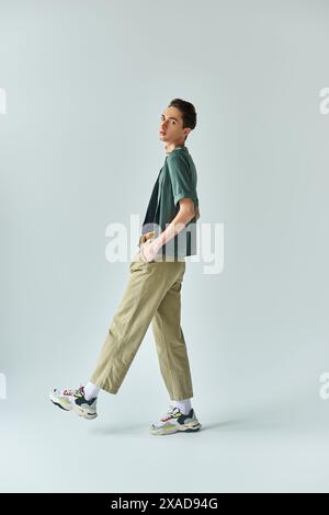 Eine junge queere Person posiert selbstbewusst in einem Studio, trägt ein stilvolles T-Shirt und Khaki-Hose auf grauem Hintergrund. Stockfoto