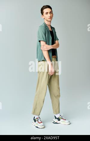 Eine junge queere Person posiert selbstbewusst in einem Studio, in einem grünen Hemd und Khaki-Hose, vor grauem Hintergrund. Stockfoto