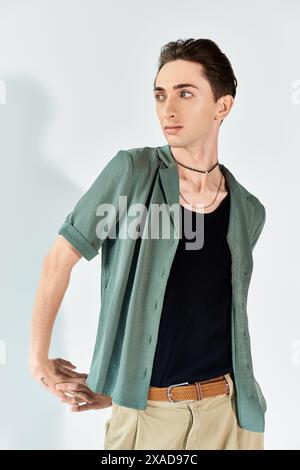 Eine junge queere Person posiert selbstbewusst in einem Studio mit einem grünen Hemd und einer braunen Hose auf grauem Hintergrund. Stockfoto