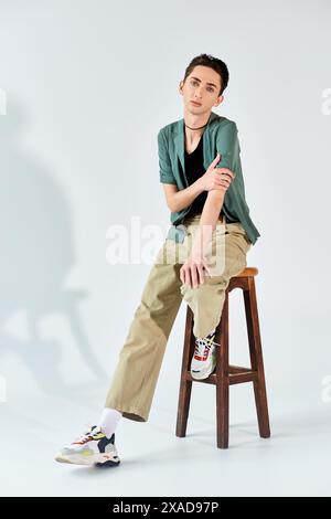 Eine junge queere Person sitzt selbstbewusst auf einem Hocker in einem Studio und strahlt Stolz und Ermächtigung vor grauem Hintergrund aus. Stockfoto