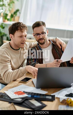 Zwei Männer, ein schwules Paar, sitzen an einem Tisch und konzentrieren sich intensiv auf einen Laptop, während sie zusammen in einer Designerwerkstatt arbeiten. Stockfoto