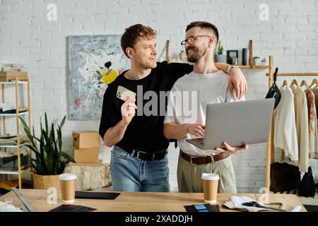 Zwei Männer, ein schwules Paar, stehen in einer Designerwerkstatt, konzentrieren sich auf einen Laptop-Bildschirm, während sie trendige Kleidung diskutieren. Stockfoto