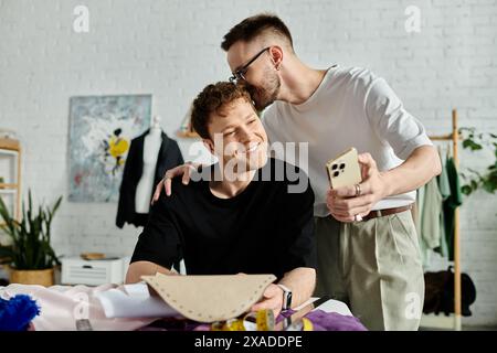 Schwuler Mann küsst den Kopf eines Freundes in einer Designer-Werkstatt, der zusammen an der Gestaltung trendiger Kleidung arbeitet. Stockfoto