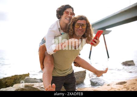 Glücklicher Mann, der seine Freundin auf dem Rücken trägt und Spaß am Strand hat Stockfoto