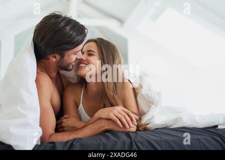 Ein Paar, lächeln und unter der Decke im Bett für morgendliche Romantik, Liebe und Bindungen am Wochenende. Beziehung, Mann und Frau, die zusammen zu Hause liegen Stockfoto