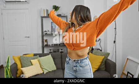 Eine fröhliche junge Frau tanzt frei in ihrem gemütlichen Wohnzimmer, das mit modernem Dekor und lebhaften Kissen gefüllt ist. Stockfoto