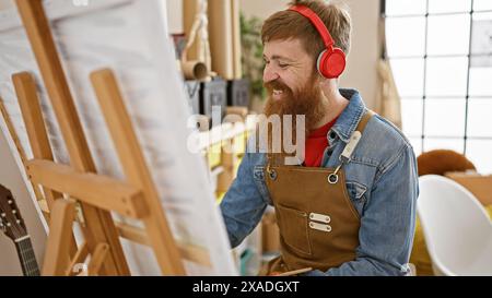 Hübscher, junger, rothaariger bärtiger Mann, freudig vertieft in eine Malstunde, lauscht Lieblingsliedern, während er an einer Staffelei steht, die in einem A zeichnet Stockfoto