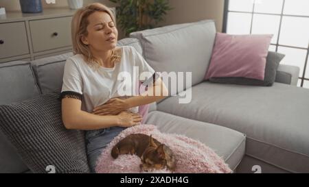 Junge blonde Erwachsene Frau in einem Wohnzimmer zu Hause mit einem chihuahua Hund auf dem Schoß, die Unannehmlichkeiten beim Sitzen auf einem Sofa empfindet. Stockfoto