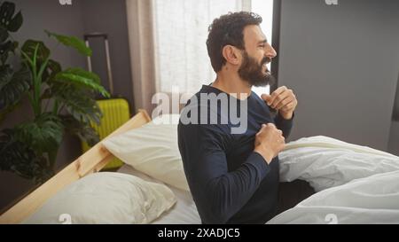 Lächelnder bärtiger Mann, der mit einem Koffer im Bett sitzt und eine Reise oder einen Umzug in ein gemütliches Schlafzimmer vorschlägt. Stockfoto