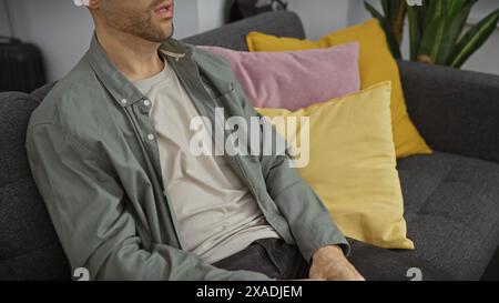 Ein bärtiger hispanischer Mann in lässiger Kleidung entspannt sich auf einem grauen Sofa, umgeben von bunten Kissen in einem modernen Wohnzimmer. Stockfoto