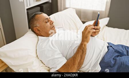 Ein reifer hispanischer Mann entspannt sich in einem Schlafzimmer, lässig gekleidet, während er sein Smartphone durchsucht. Stockfoto