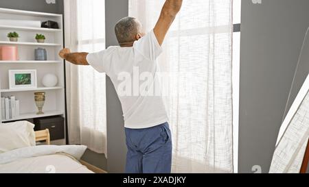 Ein hispanischer Mann mittleren Alters breitet sich am Fenster in einem gemütlichen Schlafzimmer aus und strahlt Ruhe und Komfort aus. Stockfoto