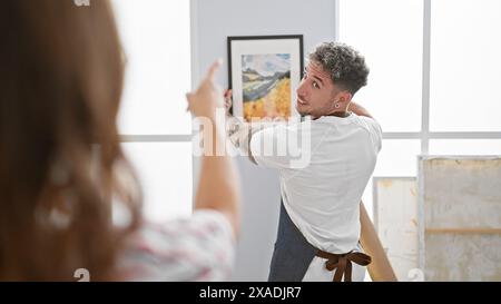 Mann und Frau arbeiten in einem hellen Kunststudio zusammen und hängen ein buntes gerahmtes Gemälde zusammen Stockfoto