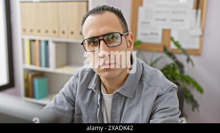 Ein selbstbewusster hispanischer Mann mit Brille und grauem Hemd posiert in einem modernen Büro. Stockfoto