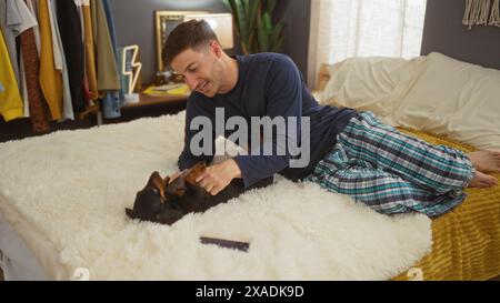 Ein junger hispanischer Mann im Pyjama liegt auf einem Bett in einem gemütlichen Schlafzimmer und spielt mit seinem Haustier-Dackelhund. Stockfoto