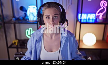 Eine junge Frau mit blonden Haaren und Kopfhörern sitzt nachts in einem lebhaften Spielzimmer. Stockfoto