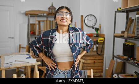 Eine junge indianerin mit langen Haaren steht in einer Tischlerei, trägt eine Schutzbrille und ein kariertes Hemd. Stockfoto