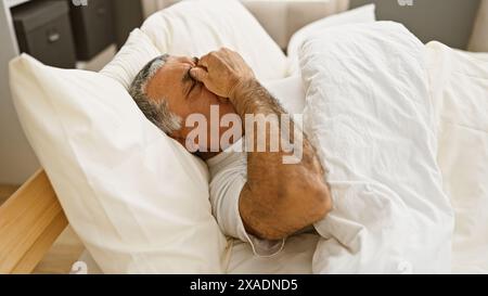 Ein Mann mittleren Alters mit grauen Haaren liegt im Bett, scheinbar verstört oder unwohl, in einem Schlafzimmer. Stockfoto