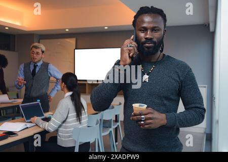 Geschäftsmann mit Dreadlocks im Büro, der am Telefon diskutiert, ernsthaft in die Kamera schaut. Er telefoniert, während seine Kollegen arbeiten. Er Stockfoto