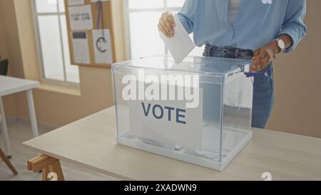 Eine junge afroamerikanische Frau gibt ihre Stimme an einer Wahlhochschule ab, wobei ihre Hände gesehen werden, wie sie einen Wahlzettel in eine transparente Box drinnen platzieren. Stockfoto