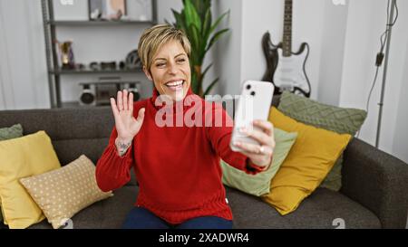 Eine lächelnde hispanische Frau mit kurzen blonden Haaren macht ein Selfie in einem gemütlichen Wohnzimmer. Stockfoto