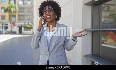 Eine schöne junge afroamerikanerin mit lockigen Haaren, die einen Blazer trägt, lächelt und spricht auf ihrem Telefon, während sie auf einer Straße in der Stadt auf einer sonnigen Sonne steht Stockfoto