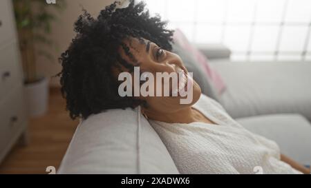 Eine wunderschöne afroamerikanische Frau mit lockigen Haaren, die lächelnd in einem stilvollen Wohnzimmer entspannen. Stockfoto