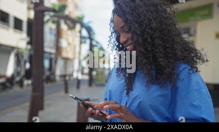Eine schöne afroamerikanische Frau mit lockigen Haaren lächelt, während sie auf einer städtischen Straße steht und ihr Telefon benutzt. Stockfoto