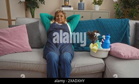 Eine junge afroamerikanische Frau mit Zöpfen, die sich in einem Wohnzimmer zu Hause entspannt, sitzt auf einem Sofa, umgeben von bunten Kissen, mit Putzgeschirr Stockfoto