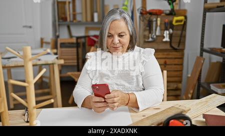 Reife Frau, die ein Smartphone in ihrem Holzbearbeitungsstudio benutzt, das mit Werkzeugen und Holz gefüllt ist Stockfoto