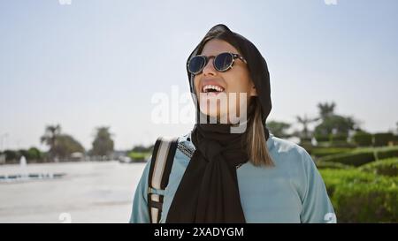 Glückliche hispanische Erwachsene Frau mit Sonnenbrille und Hijab in einer islamischen Moschee in abu dhabi, vae, symbolisiert Tourismus und kulturelle Vielfalt. Stockfoto